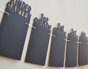 May include: Black and white paper banner with the words "Kindergarten", "1st Grade", "2nd Grade", "3rd Grade", "4th Grade", "5th Grade", and "6th Grade" cut out. The banner is strung with white thread.