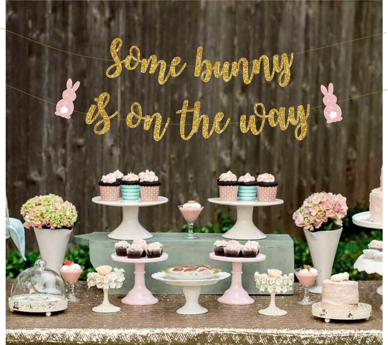 May include: A gold glitter banner with the text "Some bunny is on the way" and pink bunny cutouts. The banner is hanging above a table with cupcakes, cake stands, and floral arrangements.