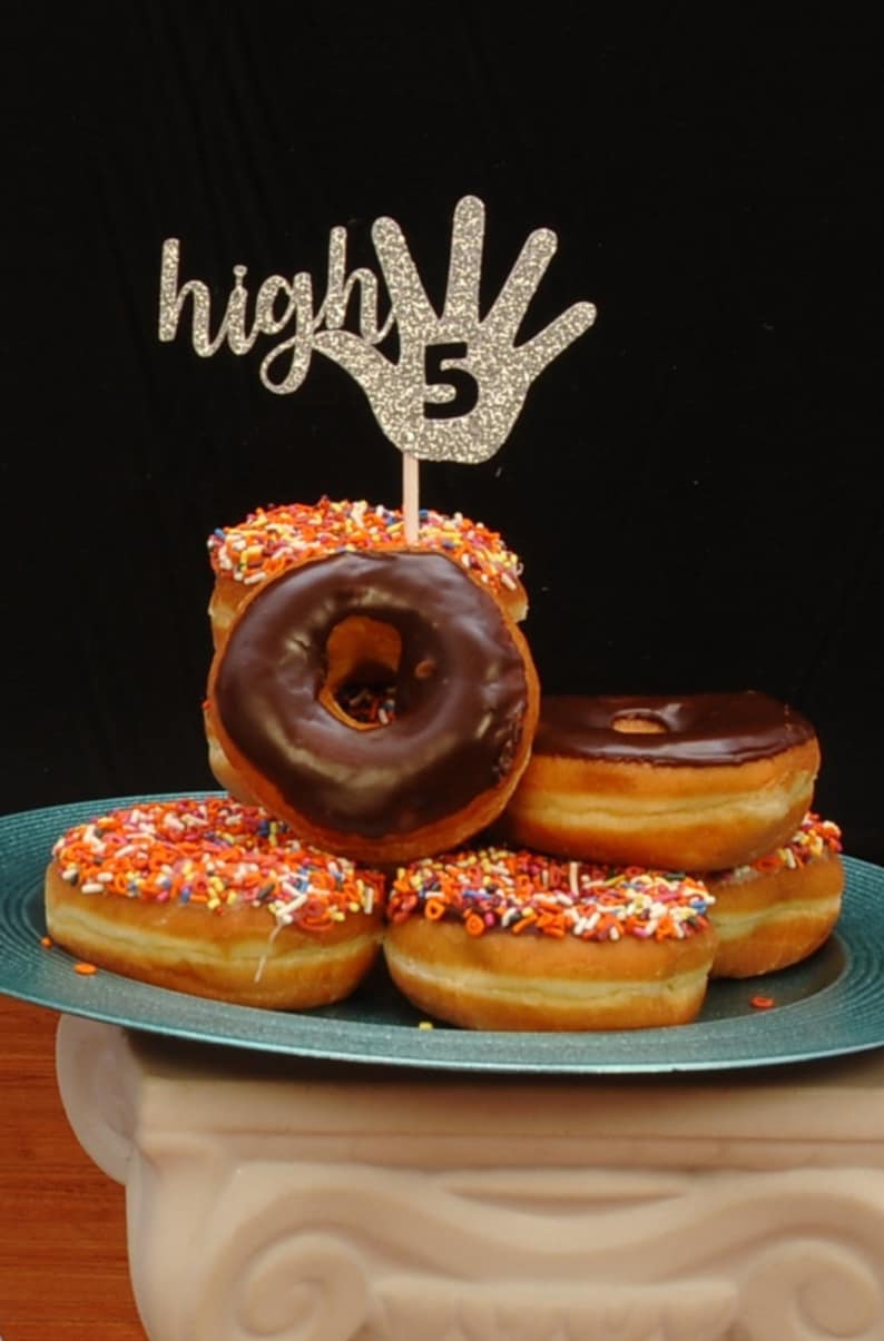 May include: A stack of chocolate frosted donuts with colorful sprinkles, topped with a silver glitter high five hand with the number 5 on it.