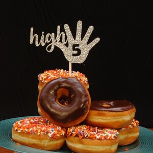 May include: A silver glitter high five hand with the number 5 on it, on a stick, is stuck into a stack of chocolate frosted donuts with colorful sprinkles. The donuts are on a teal plate.