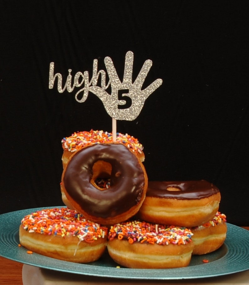 May include: A silver glitter high five hand with the number 5 on it, on a stick, is stuck into a stack of chocolate frosted donuts with colorful sprinkles. The donuts are on a teal plate.