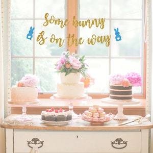 May include: A gold glitter banner with blue bunny cutouts that reads "Some bunny is on the way". The banner is hanging above a table with three cakes, cupcakes, and macarons.