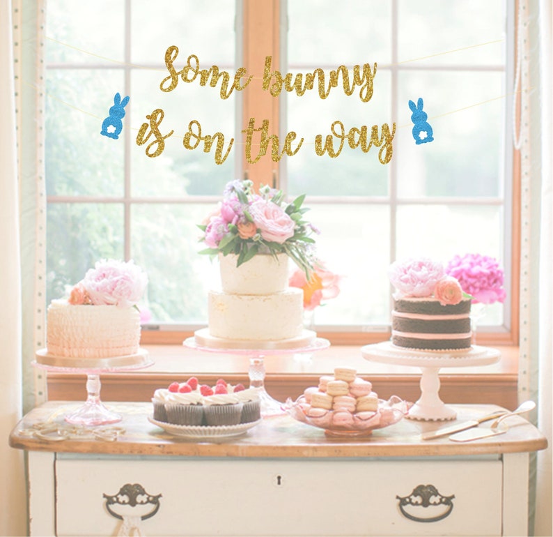 May include: A gold glitter banner with blue bunny cutouts that reads "Some bunny is on the way". The banner is hanging above a table with three cakes, cupcakes, and macarons.