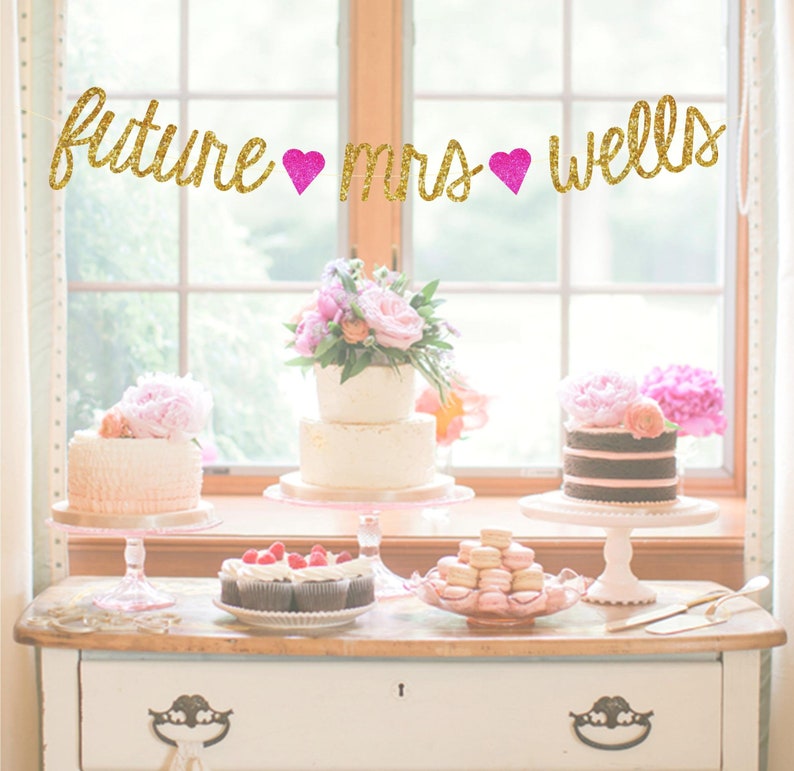 May include: A gold glitter banner with the text "future mrs wells" and pink hearts hanging above a table with three cakes and cupcakes. The cakes are decorated with flowers and frosting.