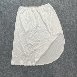 May include: White half slip made of a silky fabric. The slip has a scalloped lace trim along the hemline. The garment is laid flat on a gray surface.