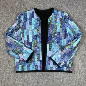 May include: A long-sleeved jacket featuring a patchwork design of vertical fabric strips in various shades of blue, teal, and purple with intricate patterns. The garment has a solid black trim along the open front, neckline, and cuffs.