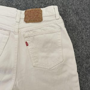 May include: Close-up of off-white denim jeans. The jeans feature a classic five-pocket design, a small red tag, and a brown leather patch with text. The jeans are made of a sturdy denim material.