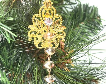 Op de afbeelding: Gouden kerstversiering met een filigraanontwerp en kristallen accenten. De versiering hangt aan een wit lint aan een groene kerstboom.