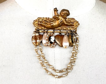 May include: A gold-toned brooch featuring a cherub with wings, surrounded by a decorative frame. The brooch has a dangling chain of pearls and charms, including hearts, a pearl drop, and a cameo.
