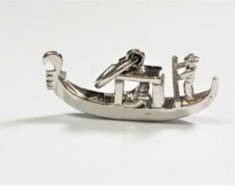 May include: A silver gondola charm with a detailed design. The charm features a gondolier, a passenger, and intricate details of the boat. The charm is suspended from a silver ring, ready to be added to a necklace or bracelet.
