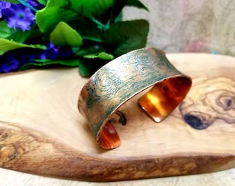 May include: A copper cuff bracelet with a textured, swirling pattern and a verdigris finish. The inside of the bracelet has a bright copper color. The bracelet is displayed on a wooden surface.