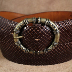 May include: A brown leather belt with a decorative, ornate, brass buckle. The belt has a snakeskin pattern.
