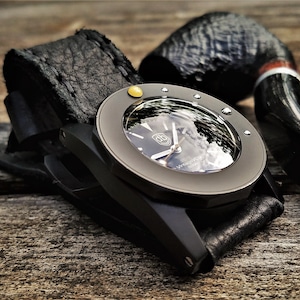 Puede incluir: Un reloj de pulsera negro con una correa de cuero negro y un acento dorado. La esfera del reloj es visible y refleja el cielo. El reloj está sobre una superficie de madera junto a una pipa negra.
