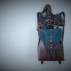 Puede incluir: Una mochila de cuero marrón con una solapa azul verdosa y una hebilla de metal decorativa. La mochila está colgada de un gancho de pared con una escultura de metal decorativa.