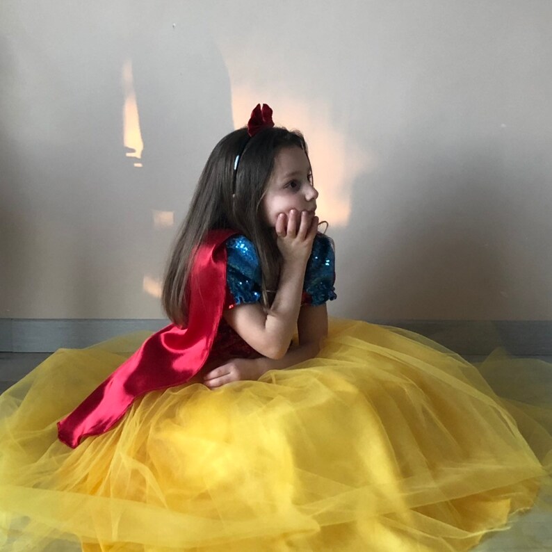 May include: A young girl dressed as Snow White, wearing a yellow tulle skirt, a red cape, and a blue and white sequined top. She is holding her chin with her hand and looking to the side.