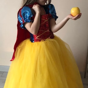 May include: A young girl dressed as Snow White in a red, blue, and yellow dress with a red cape and a yellow tulle skirt. She is holding a yellow apple in her right hand.