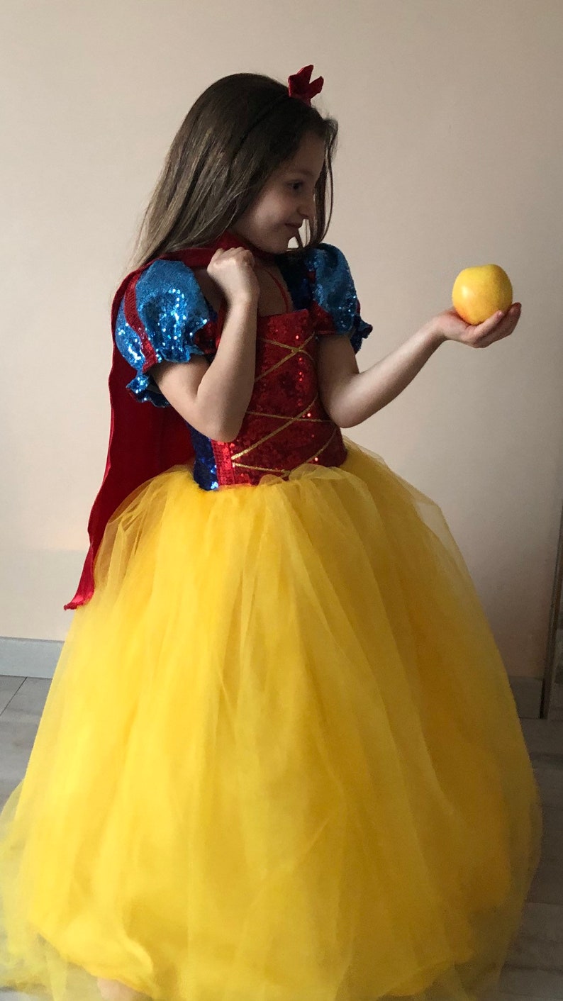 May include: A young girl dressed as Snow White in a red, blue, and yellow dress with a red cape and a yellow tulle skirt. She is holding a yellow apple in her right hand.