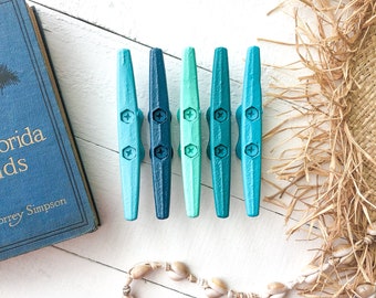 May include: Six small, wooden boat cleats painted in shades of blue and green. The cleats are arranged in a row on a white surface.