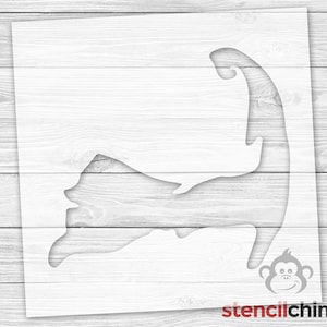 May include: A white stencil of the shape of Cape Cod, Massachusetts. The stencil is on a white wood background. The stencil is for use with paint or other mediums to create a design on a surface.