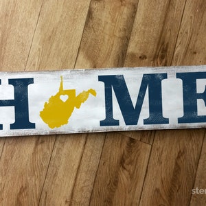 May include: A white wooden sign with the word "HOME" in blue letters. The state of West Virginia is painted in yellow with a small white heart in the center.