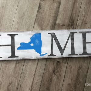 May include: White wooden sign with the word "HOME" in black lettering. The letter "O" is replaced with the outline of the state of New York in blue with a small white heart in the center.
