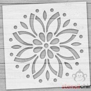May include: White stencil with a circular design featuring a large flower with multiple petals and smaller circles around the outer edge. The stencil is on a wood background.