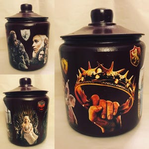 May include: A dark purple ceramic cookie jar with a decorative design featuring characters and symbols from the popular TV show Game of Thrones. The jar has a lid with a decorative knob.