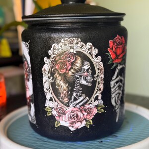 May include: Black jar with a lid, decorated with a skeleton woman design, roses, and ornate frames. The jar is sitting on a blue and white tray. The jar has a textured surface.