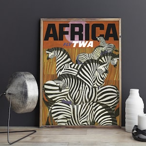 Op de afbeelding: Vintage reisposter dat TWA-vluchten naar Afrika adverteert. De poster toont een groep zebra's met zwart-witte strepen tegen een oranje-bruine achtergrond. De tekst "AFRICA FLY TWA" staat in vetgedrukte zwarte letters.
