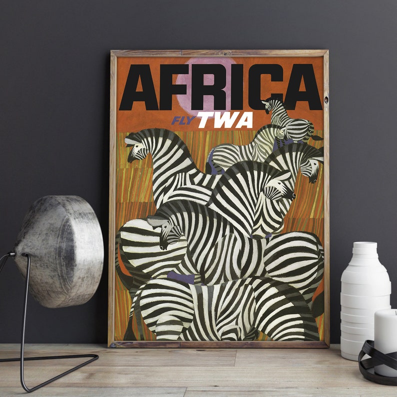 Op de afbeelding: Vintage reisposter dat TWA-vluchten naar Afrika adverteert. De poster toont een groep zebra's met zwart-witte strepen tegen een oranje-bruine achtergrond. De tekst "AFRICA FLY TWA" staat in vetgedrukte zwarte letters.