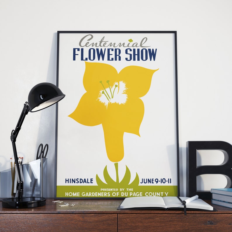 May include: A framed poster for the Centennial Flower Show, featuring a large yellow flower illustration. The poster includes the text "HINSDALE JUNE 9-10-11" and "PRESENTED BY THE HOME GARDENERS OF DU PAGE COUNTY". A black desk lamp and office supplies are on the desk.