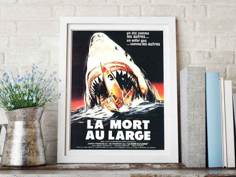 May include: A vintage movie poster for the film "Jaws 2" in French, titled "La Mort Au Large". The poster features a large great white shark with its mouth open, about to attack a small sailboat. The poster is framed in white.