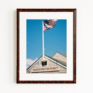 May include: A framed photograph showing the American flag waving in the wind against a clear blue sky. The photo includes a building with the text "Chatham Pier Fish Market" in red lettering.