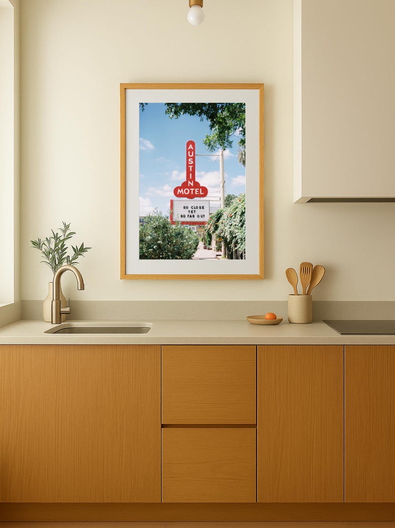 May include: A framed print featuring a vintage motel sign with the text "AUSTIN MOTEL" in red neon against a blue sky. The sign also displays the phrase "SO CLOSE YET SO FAR OUT". The print is displayed in a kitchen setting.