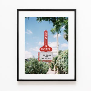 May include: A framed photograph of the Austin Motel sign against a blue sky. The sign is red with white lettering, and the text "SO CLOSE YET SO FAR OUT" is visible below. Green foliage frames the scene.
