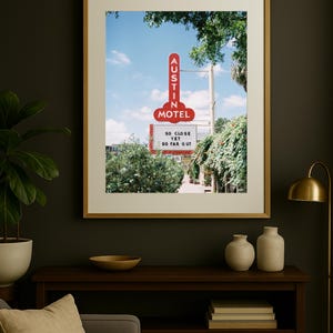 May include: A framed print featuring a vintage motel sign. The red neon sign reads "Austin Motel" against a blue sky. Below the sign, text reads "So close yet so far out." The print is displayed on a dark wall.