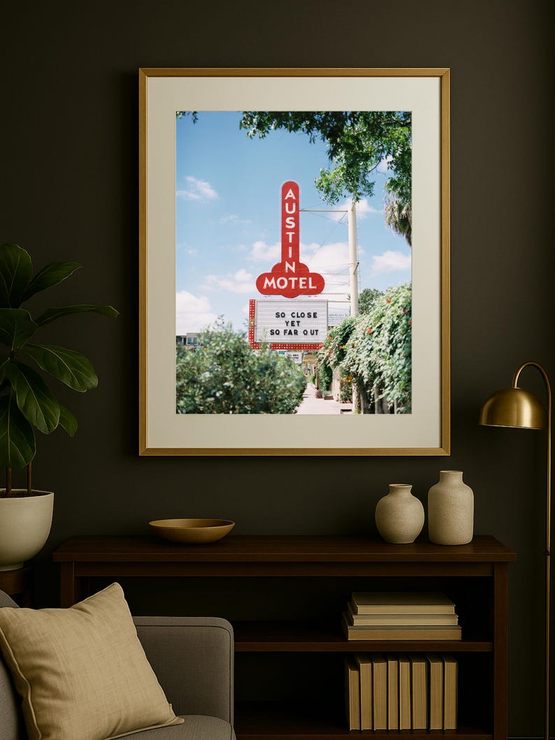 May include: A framed print featuring a vintage motel sign. The red neon sign reads "Austin Motel" against a blue sky. Below the sign, text reads "So close yet so far out." The print is displayed on a dark wall.