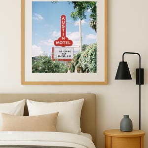 May include: A framed print featuring a vintage motel sign in Austin, Texas. The red sign reads "AUSTIN MOTEL" and "SO CLOSE YET SO FAR OUT." The image is set against a blue sky and framed in a light wood frame.