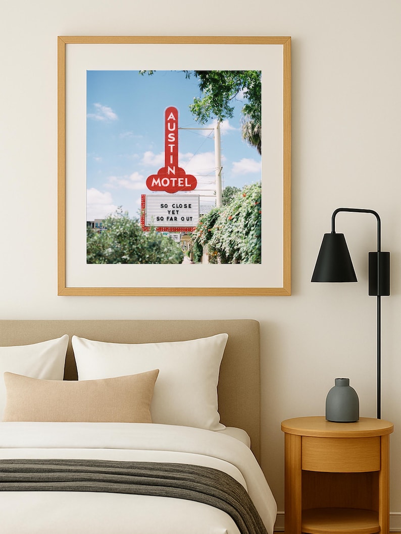 May include: A framed print featuring a vintage motel sign in Austin, Texas. The red sign reads "AUSTIN MOTEL" and "SO CLOSE YET SO FAR OUT." The image is set against a blue sky and framed in a light wood frame.