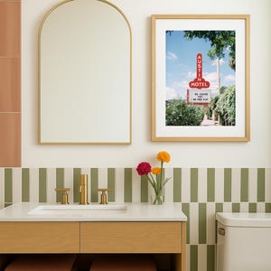 May include: A bathroom scene featuring a wooden vanity with a white countertop, gold fixtures, and a framed print of an Austin Motel sign. The walls have a green and white striped tile design, with a gold-framed mirror and a light fixture above.