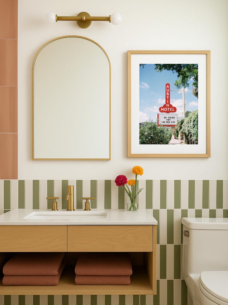 May include: A bathroom scene featuring a wooden vanity with a white countertop, gold fixtures, and a framed print of an Austin Motel sign. The walls have a green and white striped tile design, with a gold-framed mirror and a light fixture above.