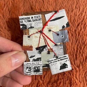 May include: A wooden board with newspaper clippings and images related to UFOs. Headlines include "Hundreds in State See 'Flying Saucers'" and "Pilots spot mile-wide UFO close to Alderney." Red thread connects the clippings.