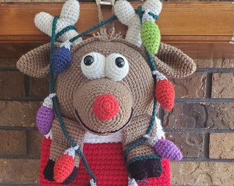 May include: A crocheted reindeer stocking with a red and white striped body and a brown reindeer head with big eyes. The stocking is decorated with a string of colorful lights.