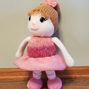 Può includere: Una bambola di lana che indossa un vestito rosa e bianco sfumato con una gonna rosa e uno chignon marrone. La bambola ha un viso bianco con occhi neri e un sorriso rosa.