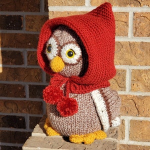 May include: A knitted owl wearing a red knitted hooded scarf. The owl is brown and white with yellow feet and black eyes. The owl is sitting on a brick surface.