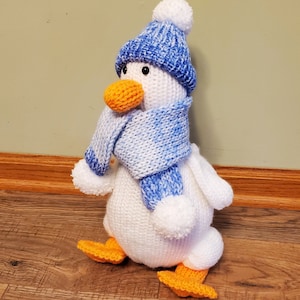 May include: White knitted penguin wearing a blue and white knit hat and scarf. The penguin has orange feet and a yellow beak.