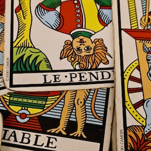May include: A tarot card titled LE PENDU featuring an upside-down figure in a yellow, red, and blue tunic, positioned above another card with the partial text IABLE. Both cards display the JODO. CAMOIN copyright mark on their white borders.