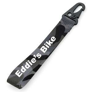 Puede incluir: Un cordón gris camuflaje con un mosquetón de metal negro. El texto "Eddie's Bike" está impreso en blanco en el cordón. Diseñado para llaves u objetos pequeños.