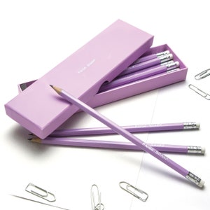 Puede incluir: Un juego de lápices morados con las palabras "Paige Sharp" impresas en ellos, en una caja morada. Los lápices están dispuestos en una fila, con los extremos de las gomas mirando hacia el espectador.