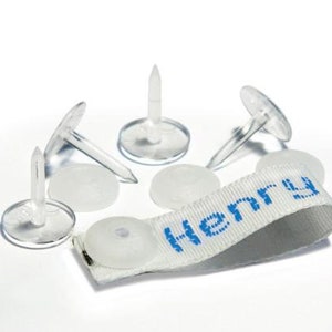May include: A collection of clear plastic name tag fasteners and a white fabric tag with the name "Henry" printed in blue. The fasteners have a flat, circular base and a pointed pin.